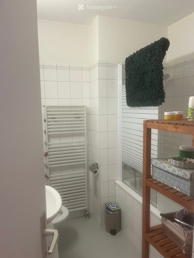A small bathroom with white tiled walls, a white radiator, a sink, a toilet paper holder, a wooden shelf with various items, a shower, and a fluffy black towel hanging on the wall.