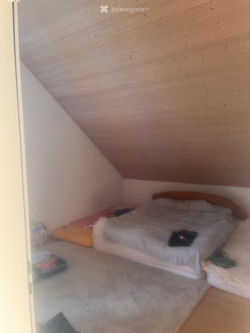 Cozy bedroom with a slanted wooden ceiling, a single bed with striped bedding, a fluffy white rug, and a storage box with folded clothes.