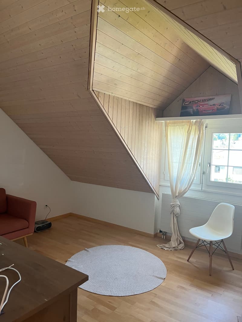 Attic room with wooden slanted ceiling, wooden floor, large window with white curtains, red sofa, white chair, round rug, wooden table, and a poster on the wall.