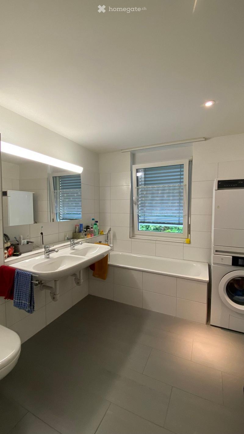 Modern bathroom with a double sink, bathtub, mirror, and washing machine. Features tiled walls, a window with blinds, and a toilet.