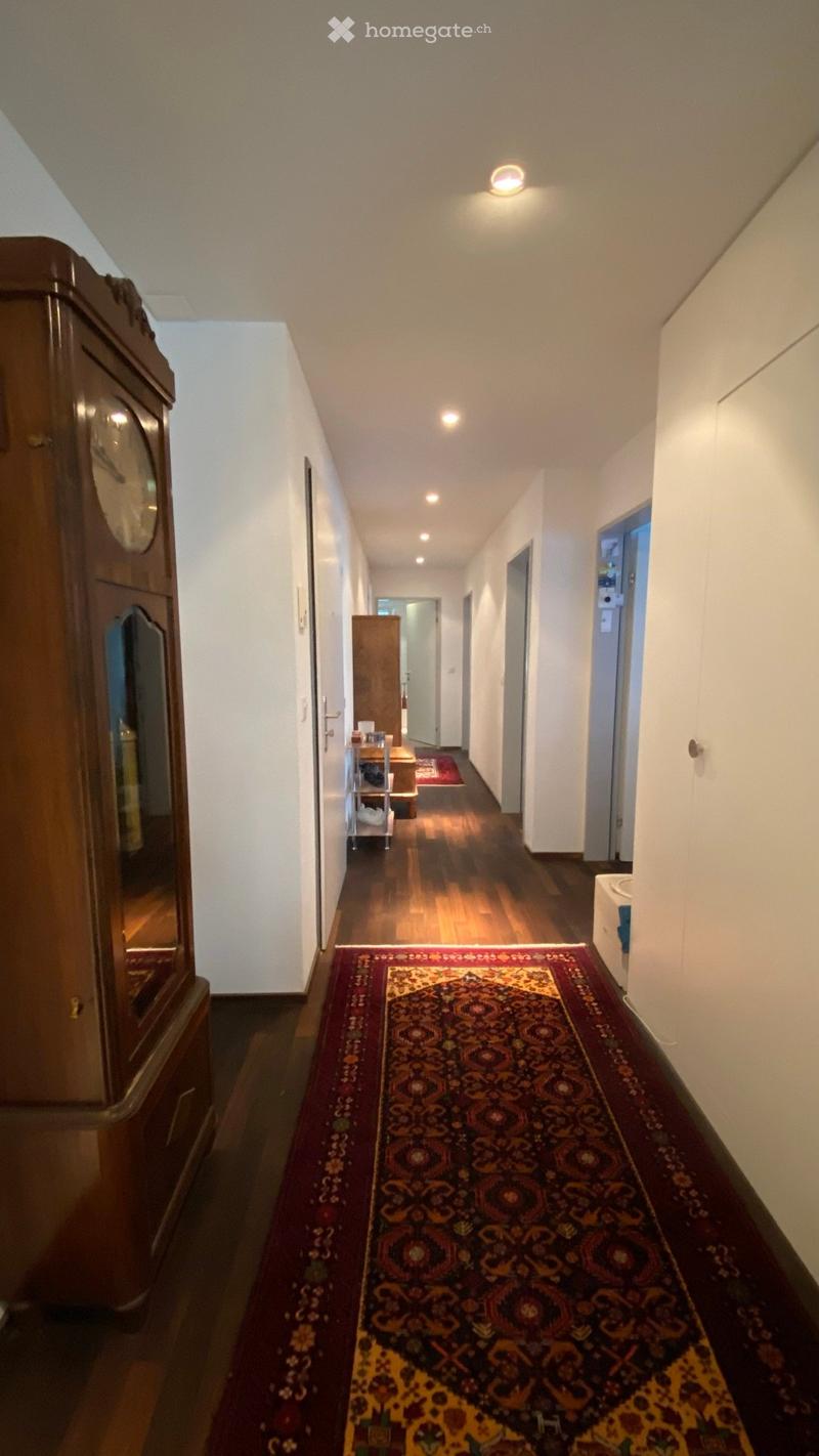 A long hallway with wooden flooring, white walls, and a patterned red and yellow runner. A wooden cabinet and a small shelf with items are visible on the left side.