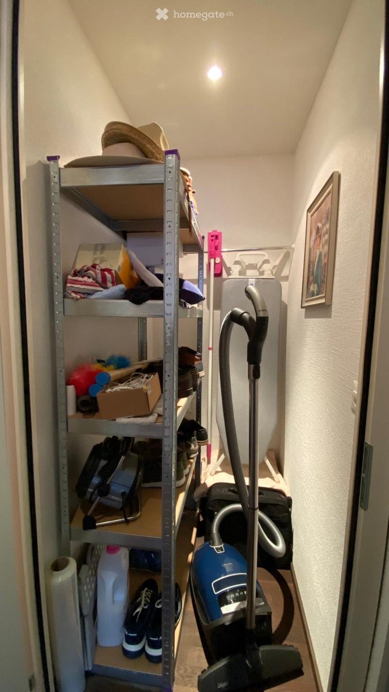 A narrow storage room with a metal shelving unit holding various items, including hats, clothes, shoes, and a vacuum cleaner on the floor.