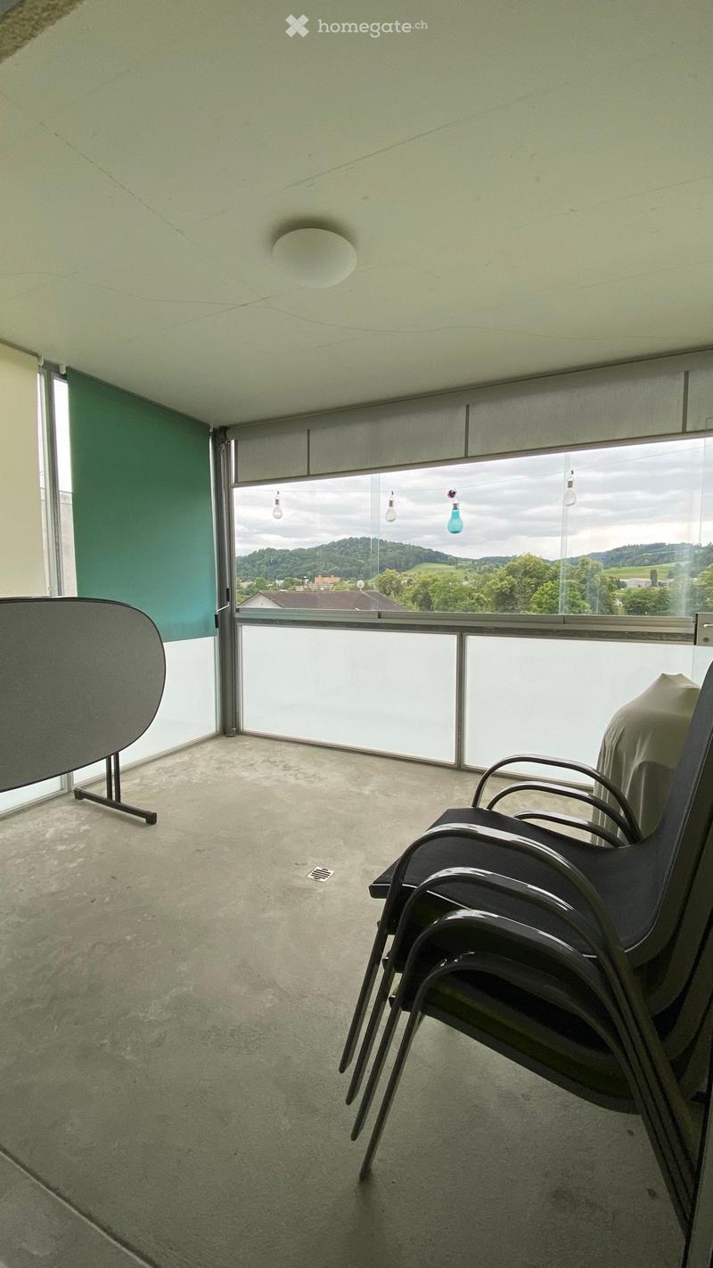 Balcony with large windows offering a scenic view, equipped with a few black chairs and a round table.