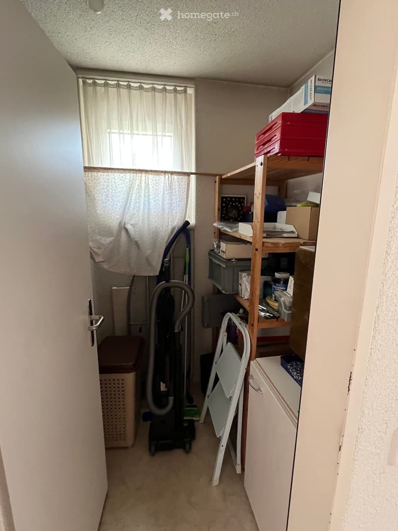 A small, organized utility or storage room with a wooden shelf holding various items, a vacuum cleaner, a folding chair, and a small cabinet.