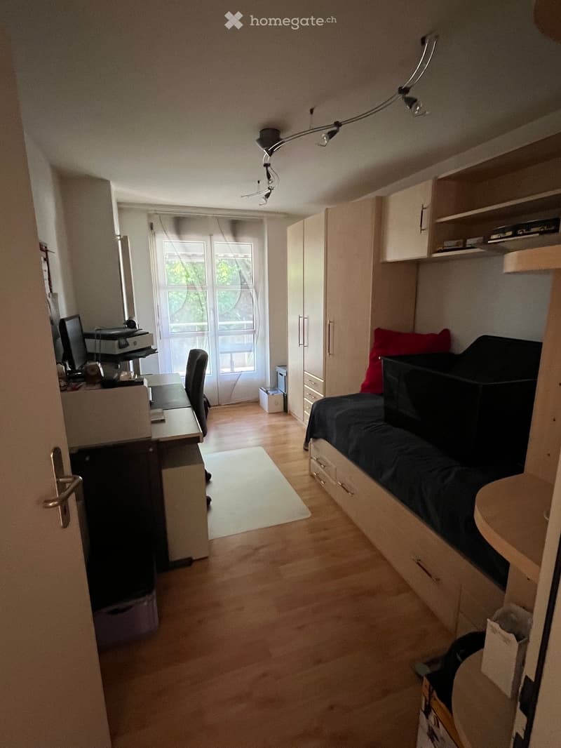 Cozy bedroom with a bed, desk, chair, computer, printer, wardrobe, and shelves. The room features wooden flooring and large windows with blinds.