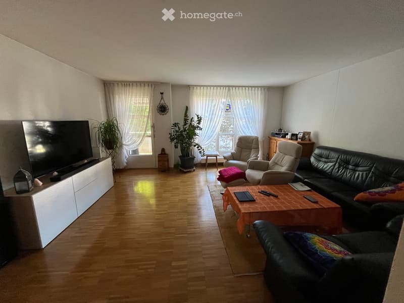 Living room with wooden flooring, large windows with white curtains, a black leather sofa, a coffee table with a red cloth, a television on a white stand, and several chairs.