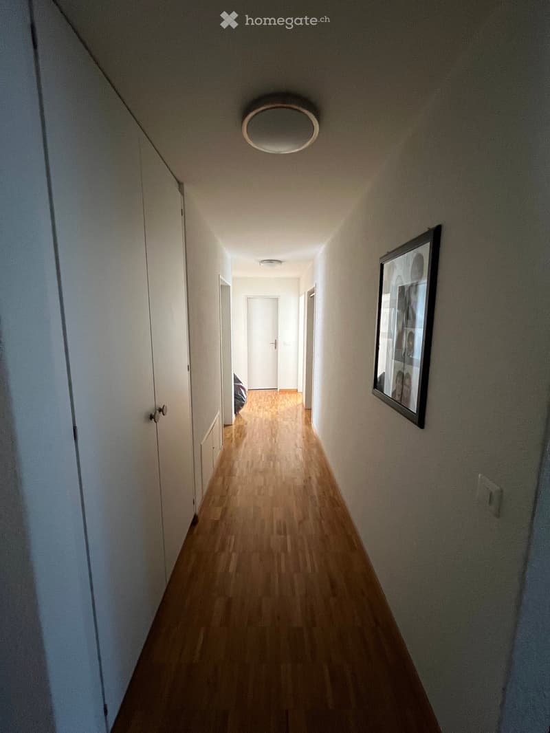 A narrow hallway with wooden flooring, white walls, and recessed lighting. A framed picture hangs on the right wall, and several doors are visible along the corridor.