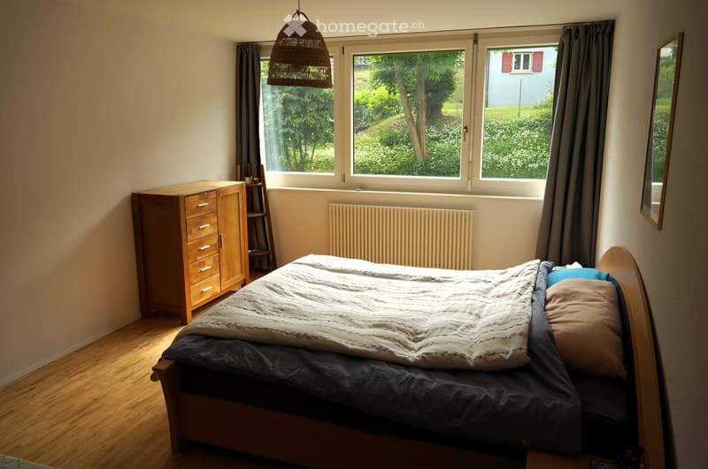 Cozy bedroom with wooden floor, large window with view of greenery, bed with pillows and blanket, wardrobe, radiator, and hanging lamp.