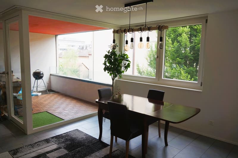 Bright dining area with a wooden dining table, four chairs, pendant lights, and a rug. Large windows offer a view of greenery, and a sliding door leads to a small outdoor space with a barbecue and artificial grass.