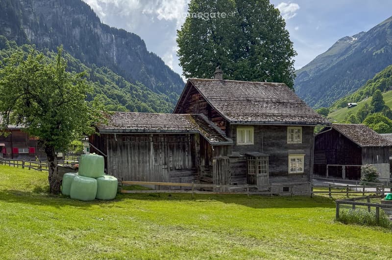 Wooden alpine house, stone chimney, wooden fence, green hay bales, mountains, grass field