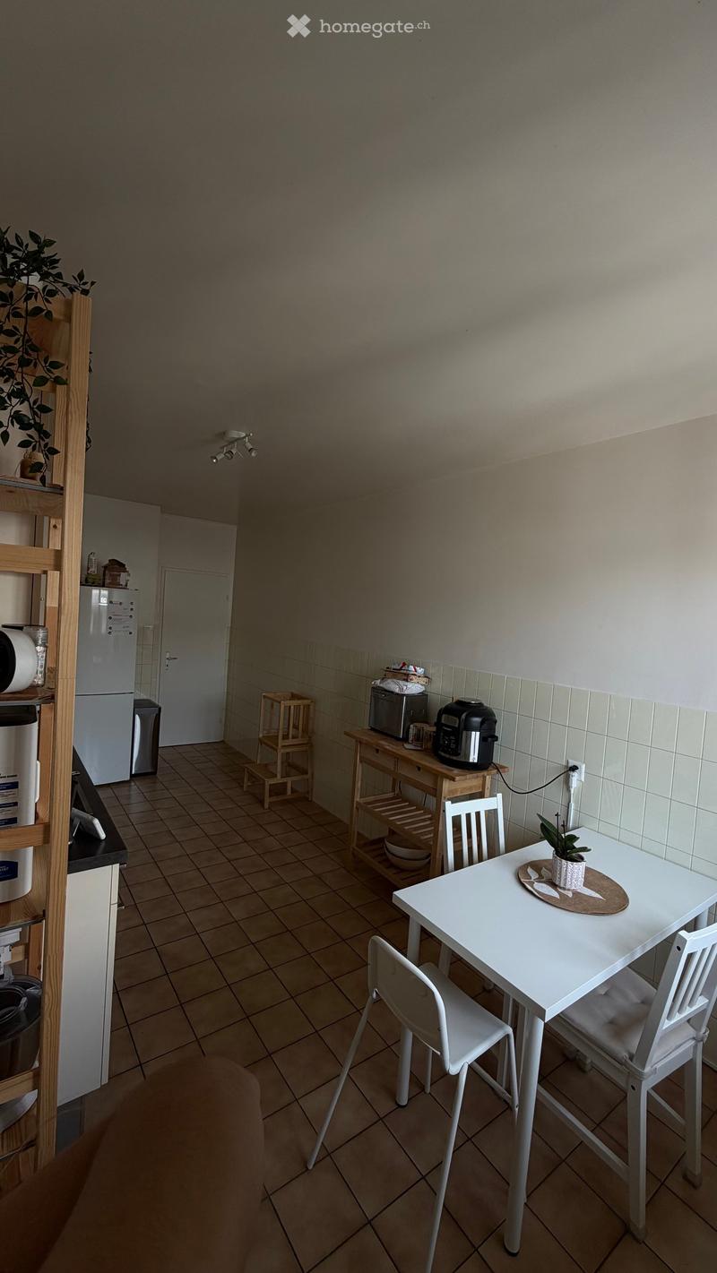 A small, modern kitchen area with tiled flooring, a white dining table with two chairs, a small wooden stool, a refrigerator, and a wooden shelf with various items.