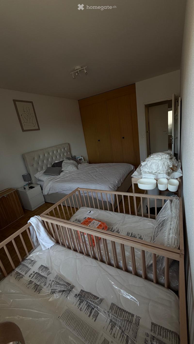 A well-lit bedroom with a large double bed, a wooden crib, a nightstand with a lamp, a wardrobe, and a picture frame on the wall.