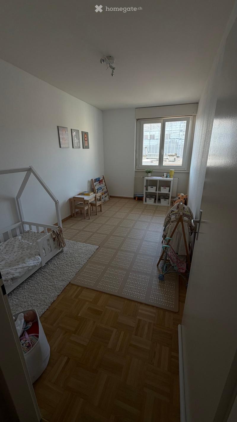 A well-lit children's room with wooden flooring, a baby bed, a small table and chairs, a shelf with books and toys, and a baby stroller near the door.