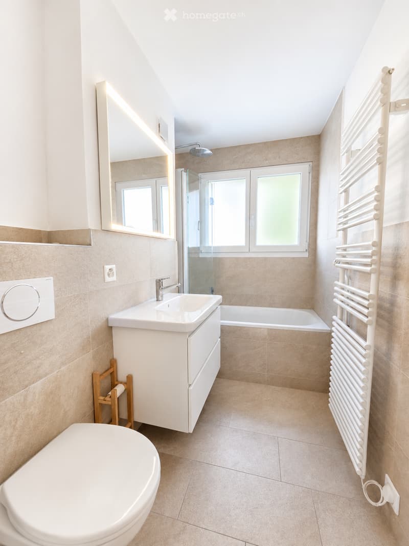 Modern bathroom with beige tiles, white fixtures, a large mirror, a bathtub, a toilet, a sink, and a radiator.