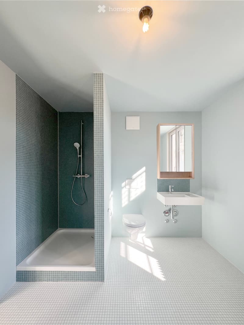 Modern bathroom with a bathtub, toilet, sink, mirror, and showerhead. Light blue tiles and white grout, with a sleek design and natural light.