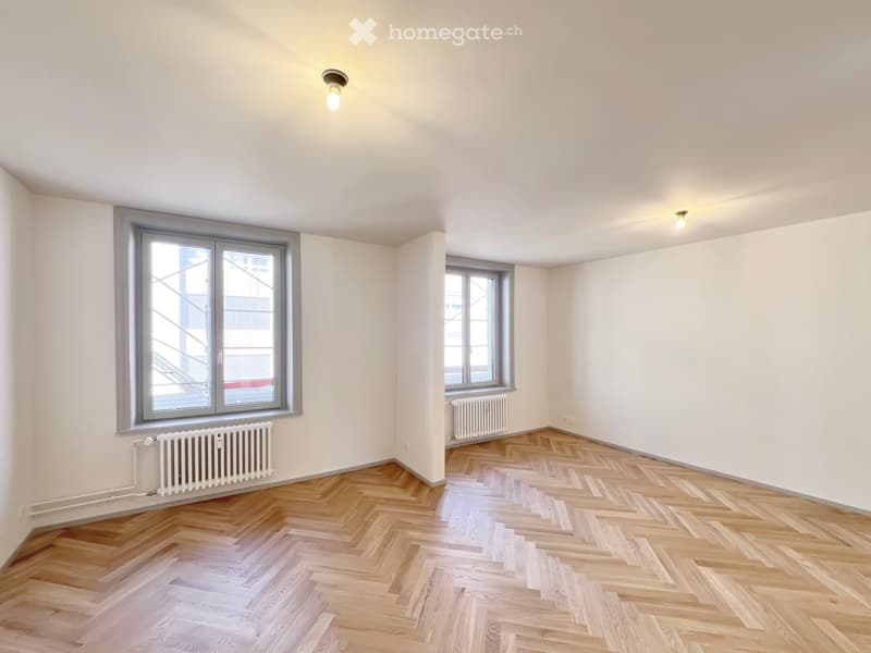 Empty room with wooden herringbone flooring, white walls, two windows, and radiators.