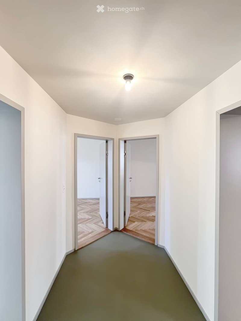 Empty hallway with light-colored walls, green carpeted floor, and two open doors leading to rooms with wooden flooring.