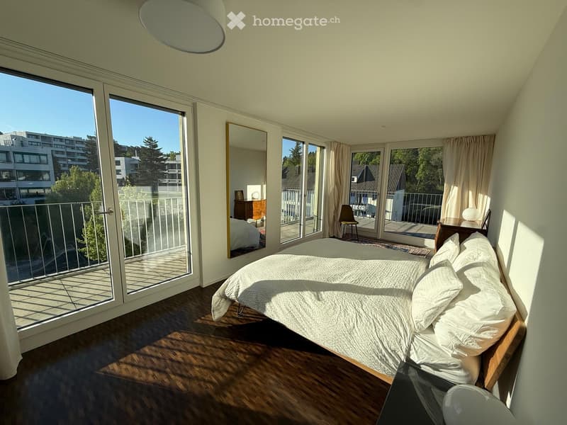 Bedroom. Bright bedroom with wooden flooring, large sliding glass doors leading to a balcony, a neatly made bed, a mirror, a chair, and a small table with a lamp.