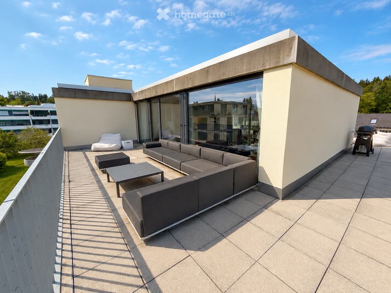 Balcony / Terrace. Modern rooftop terrace with grey sectional sofa, coffee table, ottoman, bean bag chair, and grill, surrounded by glass walls and a clear blue sky.