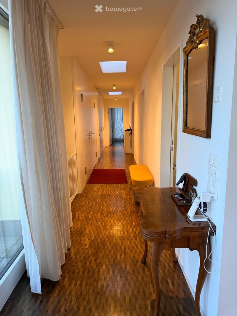 Corridor / Hallway. A narrow hallway with wooden flooring, white walls, and several doors. Features include a small red rug, a yellow bench, a wooden console table with a mirror, and a decorative mirror on the wall.