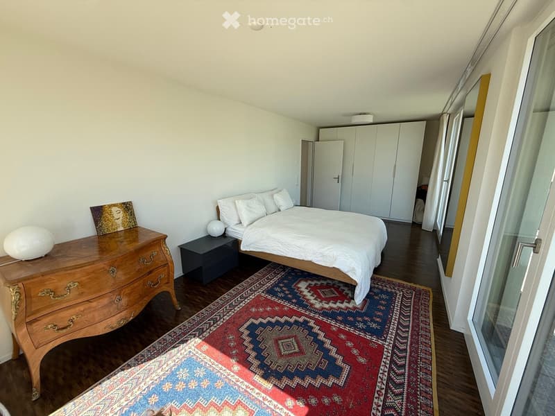 Bedroom. Modern bedroom with a large bed, nightstand, dresser, and a colorful rug. The room features wooden flooring and large windows with white curtains.