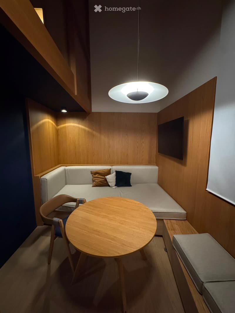 Compact living space with a sofa, wooden table, chair, pendant light, and a screen mounted on the wall.