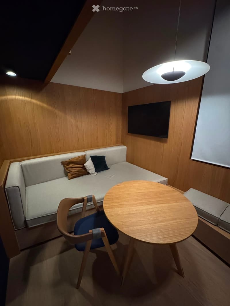 Modern, compact living space with a sofa, small round table, chair, and mounted television. The room features wooden walls and a sleek, contemporary design.