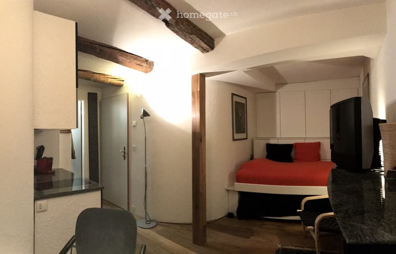Cozy bedroom with a bed, red bedding, a chair, a table, a mirror, a floor lamp, and a television. The room features exposed wooden beams and a small seating area.
