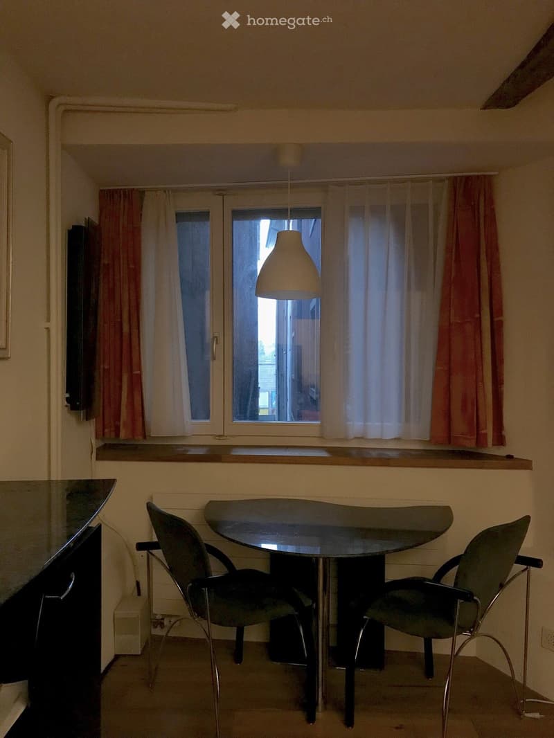 A small, furnished room with a glass table and two chairs, a window with curtains, and a hanging lamp.