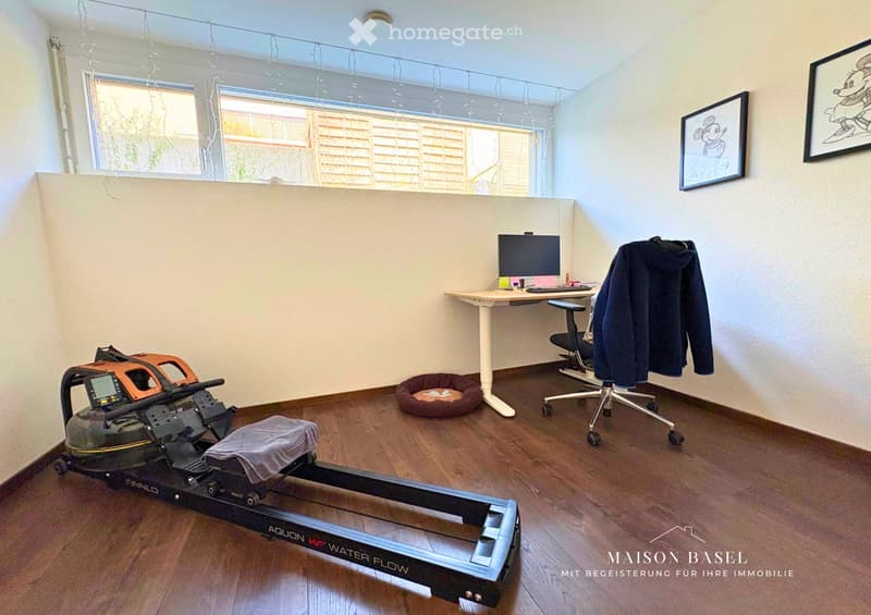 Well-lit room with wooden flooring, a desk with a computer, a chair, a dog bed, and an exercise machine. Two framed pictures on the wall and a coat hanging on the side.