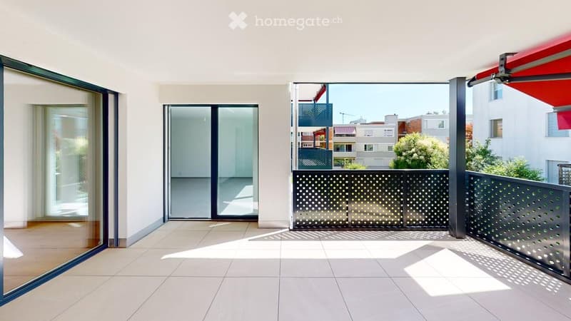 Modern balcony with large glass sliding doors, tiled floor, metal balustrade with perforated design, and a red awning partially visible.