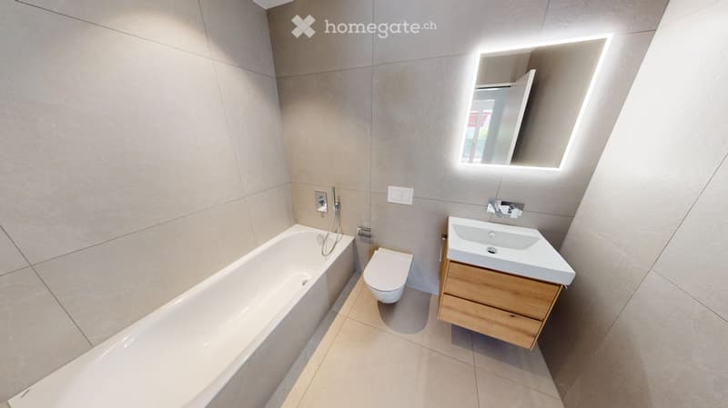 Modern bathroom with a bathtub, toilet, sink, and a wooden cabinet. Features a large mirror with built-in lighting and sleek, contemporary design.