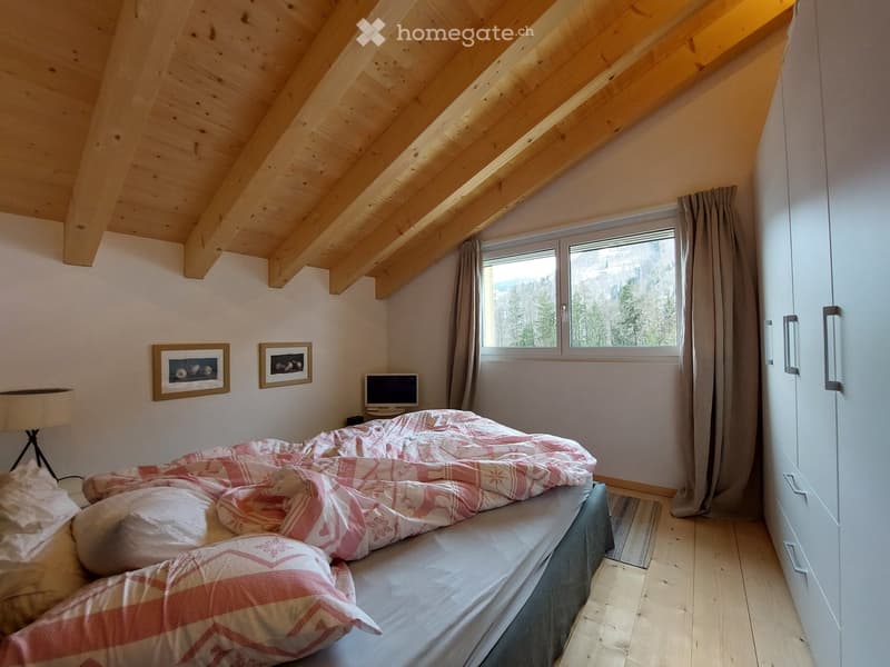 Cozy attic bedroom with wooden slanted ceilings, a bed with a red and white checkered duvet, a laptop on a nightstand, and a large window offering a scenic mountain view.