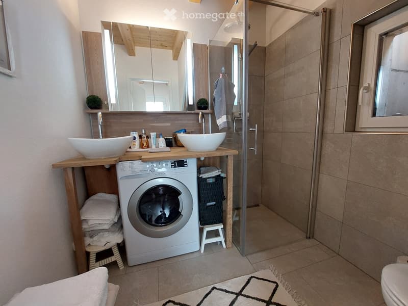Modern bathroom with a wooden vanity, two sinks, a washing machine, a shower, and a toilet. The vanity has a mirror above it and a small stool next to the washing machine.