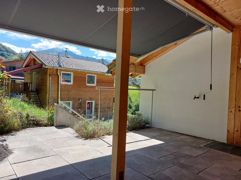 A covered outdoor patio area with wooden beams, stone tiles, and a view of a wooden house and mountains in the background.