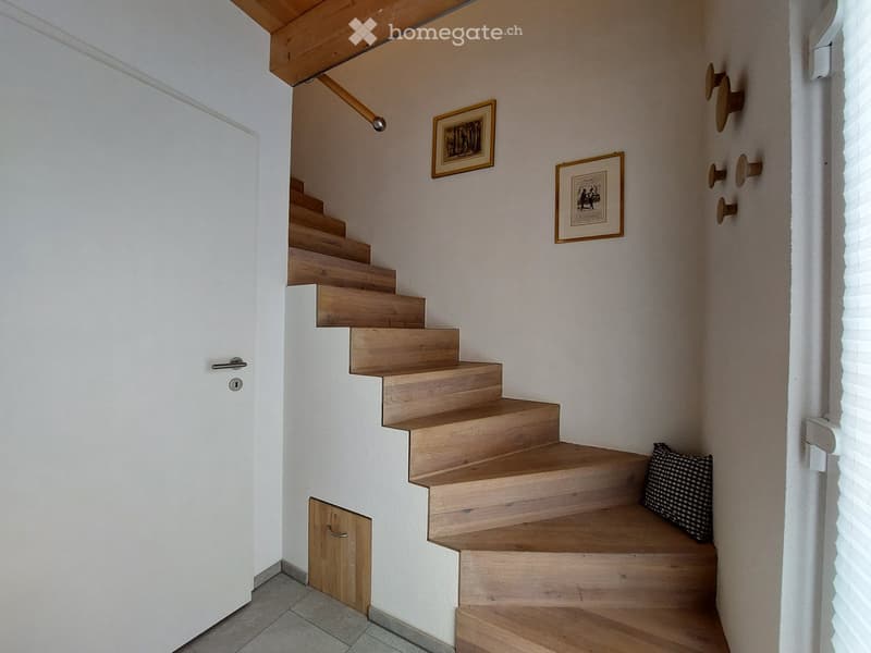 Wooden staircase with white walls, framed artwork, and a small cushioned seat at the bottom.
