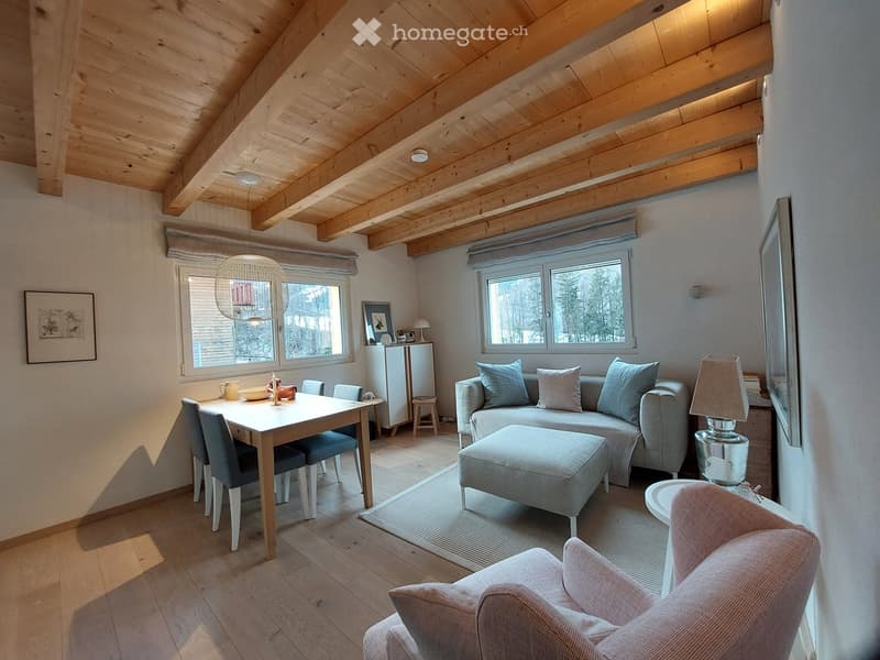 Bright living room with wooden ceiling beams, large windows, a grey sectional sofa, ottoman, dining table with chairs, and a small refrigerator.