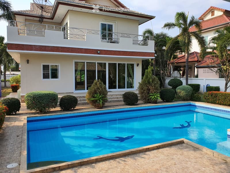 Two-story house with a swimming pool in the foreground, surrounded by trimmed bushes and palm trees. The house features a balcony on the upper floor and large windows.