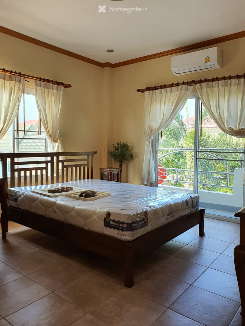Bright bedroom with a large bed, nightstand, and balcony access. The room features light-colored curtains and a view of greenery outside.