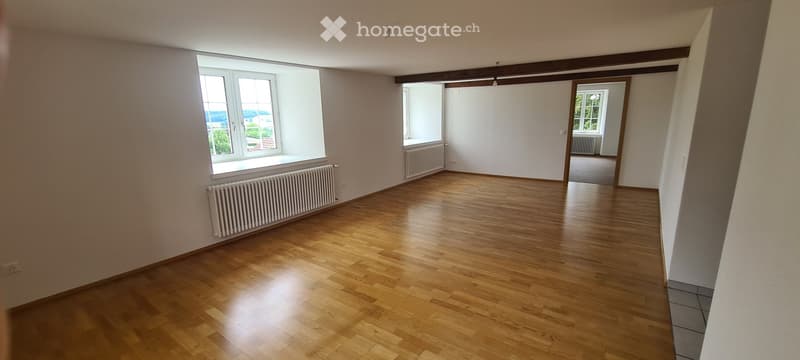 Empty room with wooden flooring, white walls, large windows, radiators, and a visible doorway leading to another room.