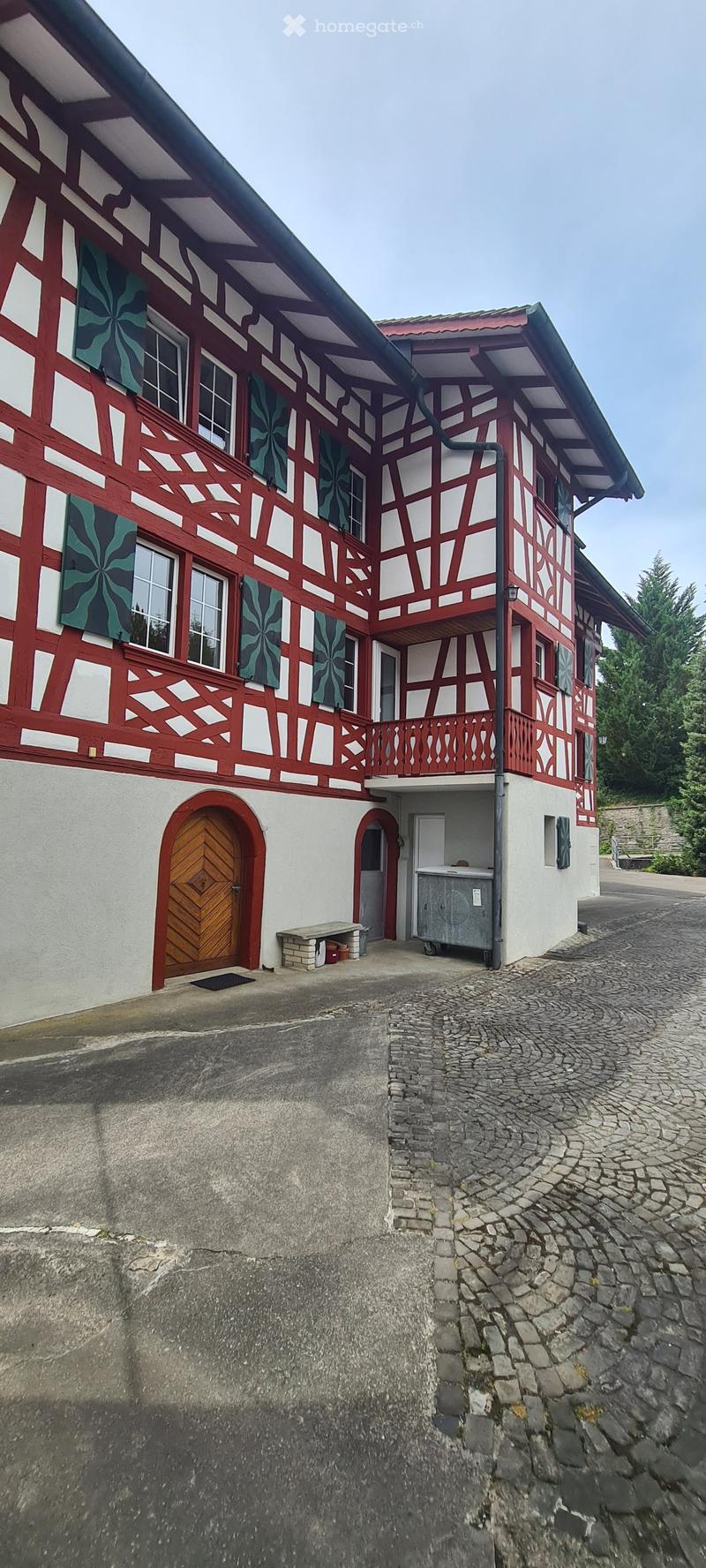 Traditional two-story house with red and white timber framing, multiple windows, and a balcony. Features a wooden front door and a small stone bench near the entrance.