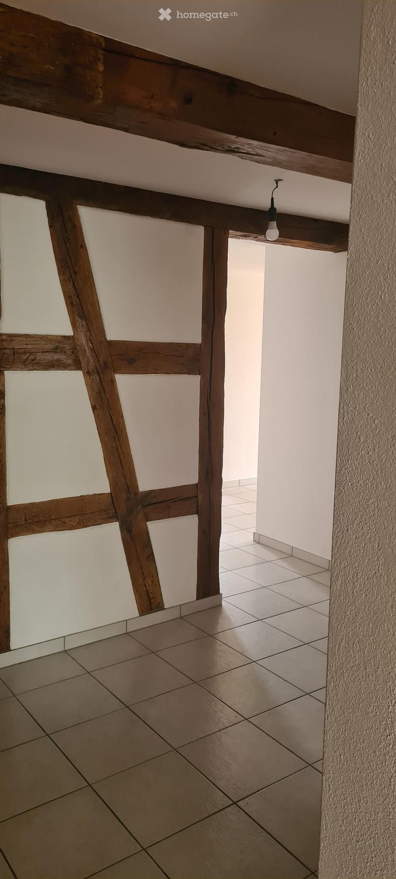 Empty room with exposed wooden beams, tiled floor, and white walls. No furniture or equipment present.