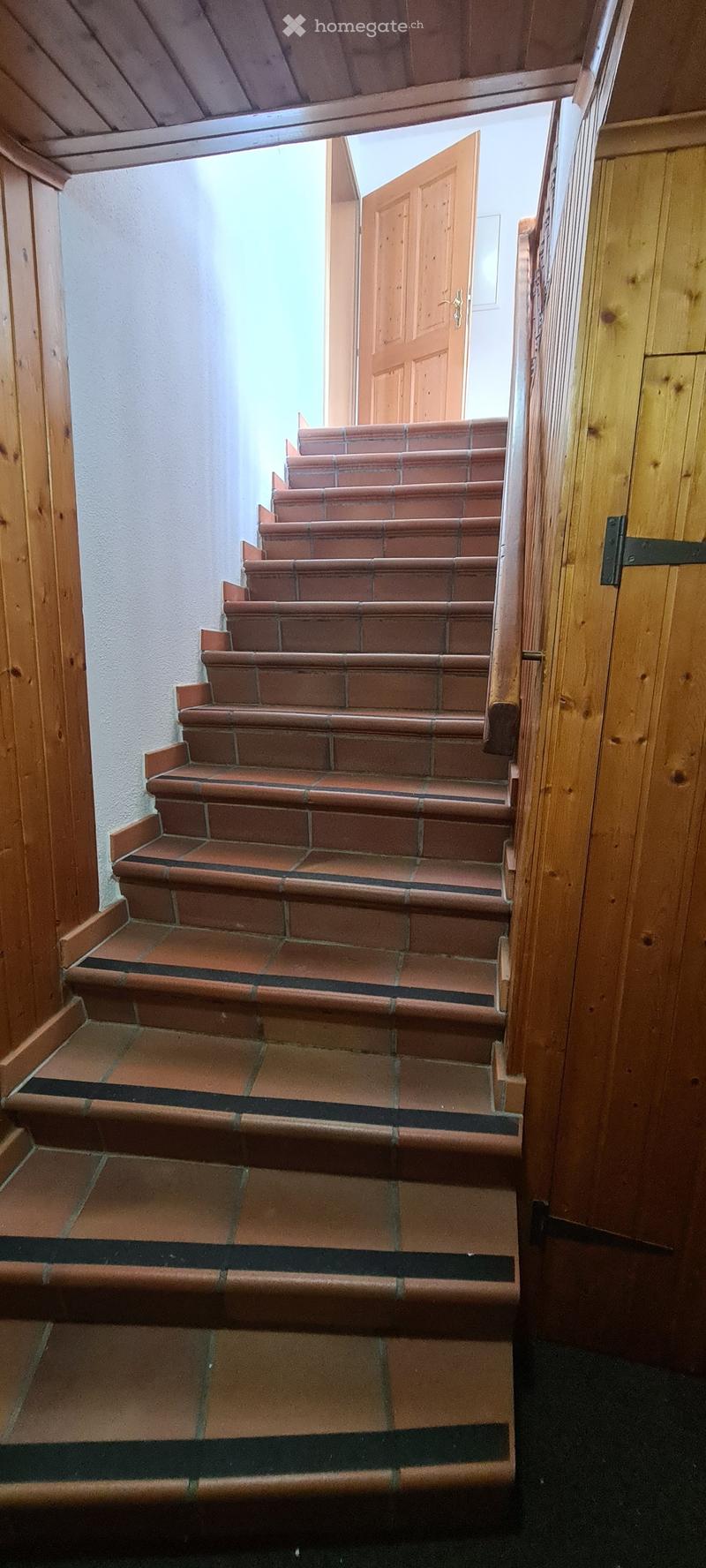 A wooden staircase with red-tiled steps and a wooden handrail, leading upwards with a wooden door at the top.