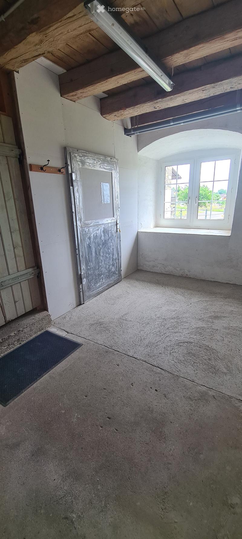 Empty room with exposed wooden beams, concrete floor, and a single window with white frame. There is a door and a small rug near the entrance.