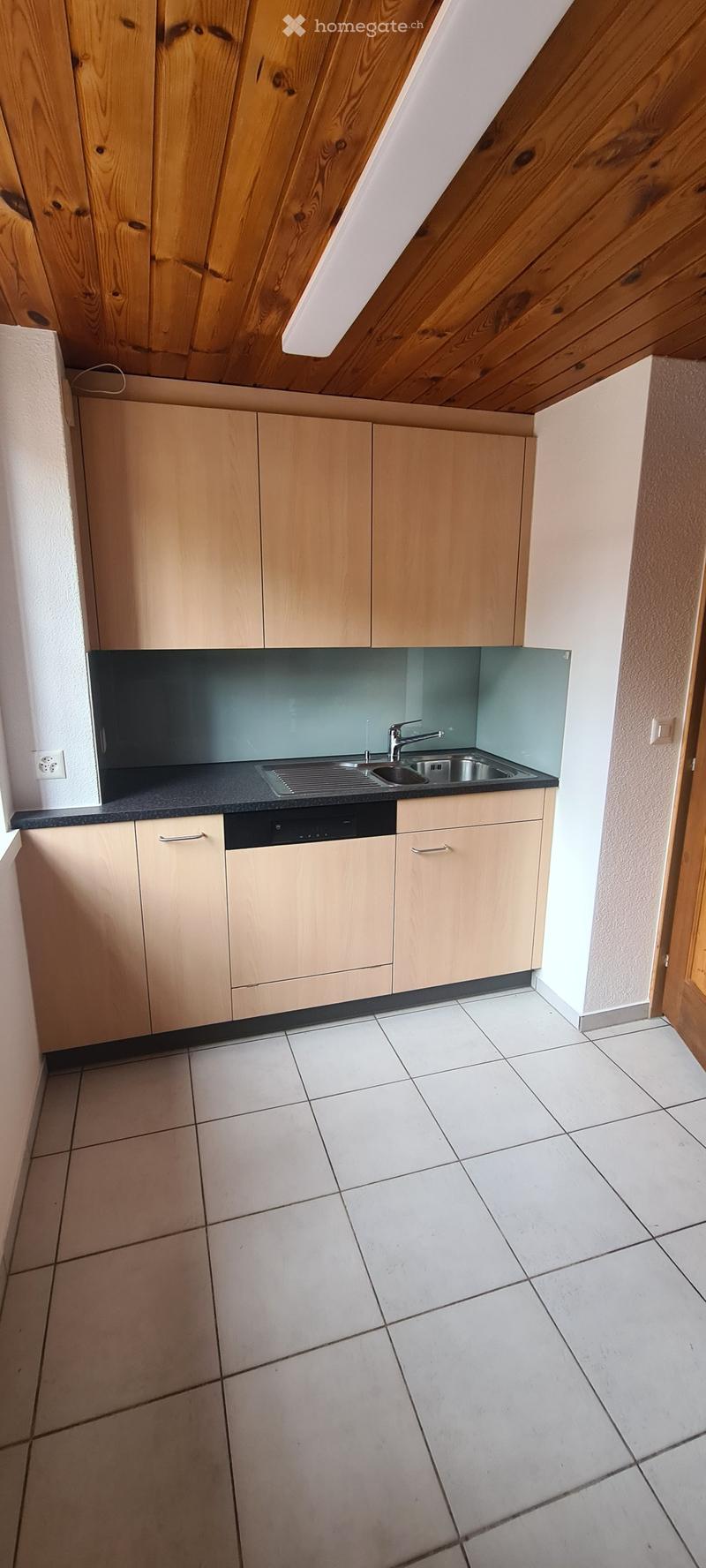 Modern kitchen with light wooden cabinets, a black countertop, a stainless steel sink, and a built-in dishwasher. The floor is tiled, and the backsplash is a sleek, dark blue glass.
