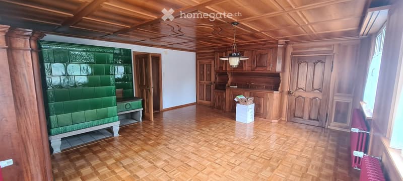 Empty room with wooden floors, wooden ceiling, and green tiled stove. No furniture or equipment present.