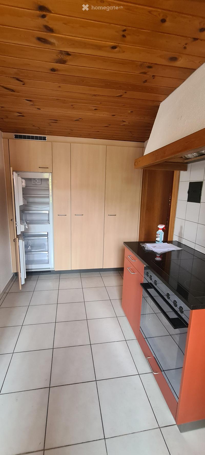 Modern kitchen with light-colored cabinets, a stainless steel refrigerator, and a black countertop with an electric stove. The floor is tiled, and the ceiling has wooden paneling.