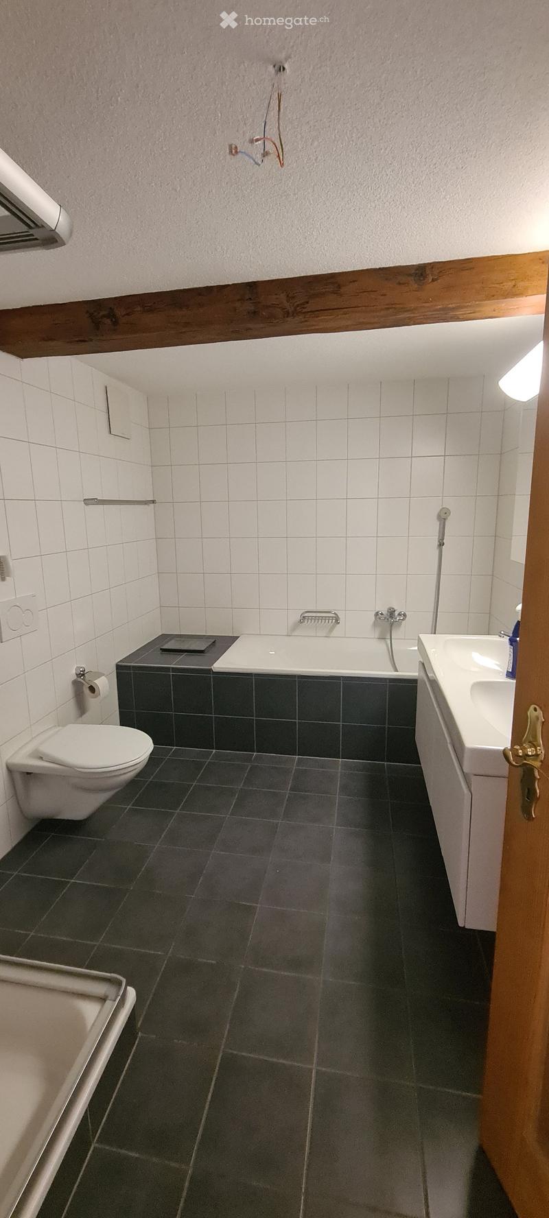 Modern bathroom with dark tiled floor, white tiled walls, bathtub, toilet, sink, and a wooden ceiling beam.