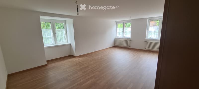 Empty room with wooden flooring, white walls, and multiple windows with white frames and curtains. Radiators are visible under the windows on the right side.
