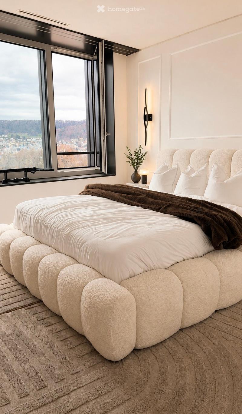 Modern bedroom with a large window offering a scenic view, a round bed with white bedding and a brown throw, a nightstand with a lamp, and a decorative vase on the windowsill.